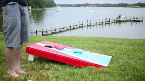 Legs of barefoot man playing bean-bag toss game near lake Stock Footage 62139982