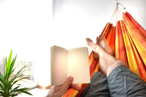 Legs of man lying down in indoor hammock holding empty notice book in his han Stock Photos