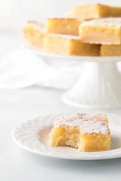 A lemon square with a bite out, with more on a dessert stand in behind. Stock Photos