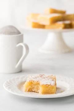 A lemon square with a bite out on a plate and desert stand with more in behind. Stock Photos