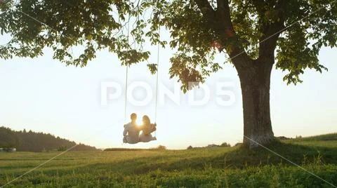 LENS FLARE SILHOUETTE: Setting sun shining over young couples shoulders on swing Stock Photos