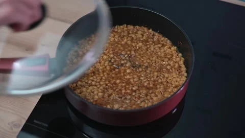 Lentils are boiling in a red pan on an induction stove. A hand places a lid on 스톡 동영상 337112023