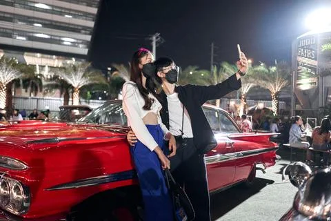 Lesbian couple taking a selfie while embracing at a night fair with antique cars Stock Photos