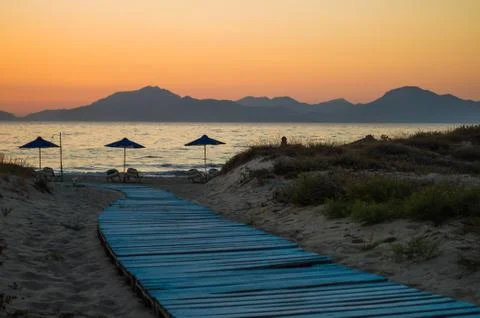 A light blue boardwalk leads down to the umbrellas on the beach at sunset Stock Photos
