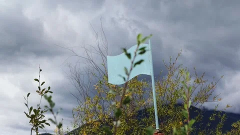 Light blue flag flutters in wind against dark clouds and distant mountains Stock Footage 344323388