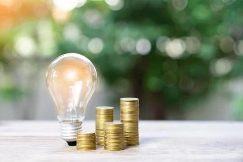 Light bulb and coins stack on out of focus plant background. Stock Photos