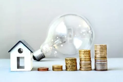 Light bulb and small house with growing stacks of coins, rising energy costs Stock Photos