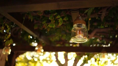 Light bulb in a transparent lamp hangs from the ceiling of a pergola in the sun Stock Footage 277083827