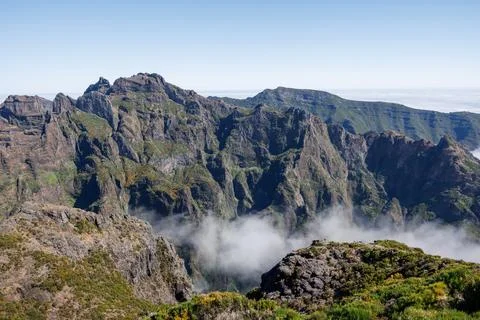 Light clouds between sharp volcanic rocks covered with shrubs on the summit.. 库存照片