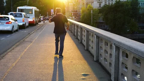 In the light of the setting sun, a man walks across a bridge and talks on the Stock Footage 256198190
