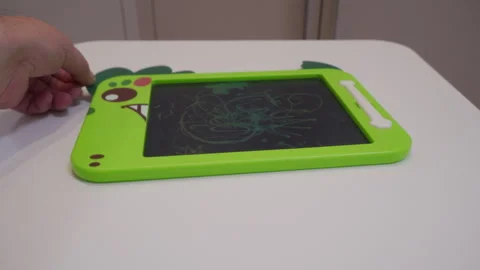 Light Thud as Hand Sets Green Dinosaur Toy Drawing Tablet on White Table Stock Footage 321268092