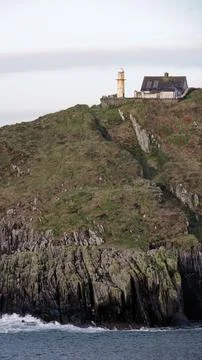 A lighthouse and a small house stand atop a steep, grassy and rocky cliff Fotos de archivo