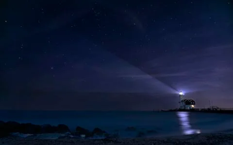 Lighthouse shining beam of light into the night sky over blue lake as beacon Stock Photos
