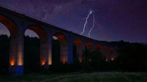 Lightning strikes above a stone bridge in the night sky with stars in the b.. Stock Photos