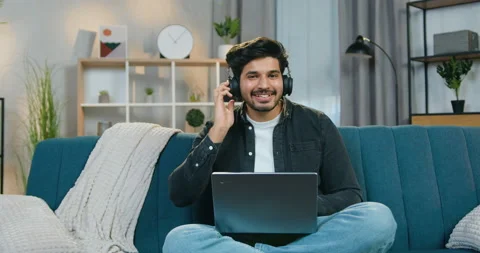 Likable joyful young bearded guy in earphones sitting on the sofa at home and Stock Footage 150156355