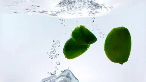 Lime fruit and ice cubes splash falling down into white cocktail and water Видео 76240741