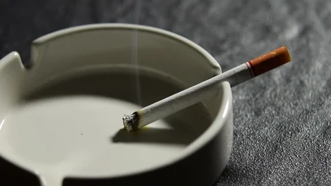 Lit cigarette lying on an empty white ashtray . Stockbeeldmateriaal 125544601