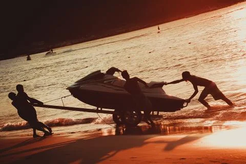 Littered horizon. Men at sunset pull hydrocycle out of the water on a sandy b Stock Photos