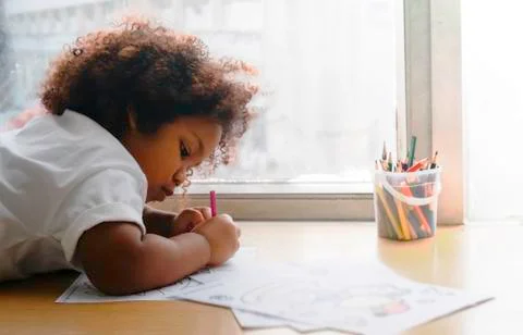 Little African girl draws and paints draws and writes lying on the floor near Stock Photos