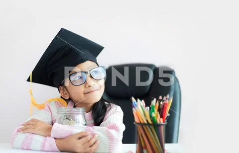 Little Asian girl wearing graduate hat hugging clear glass jar piggy bank and Stock Photos