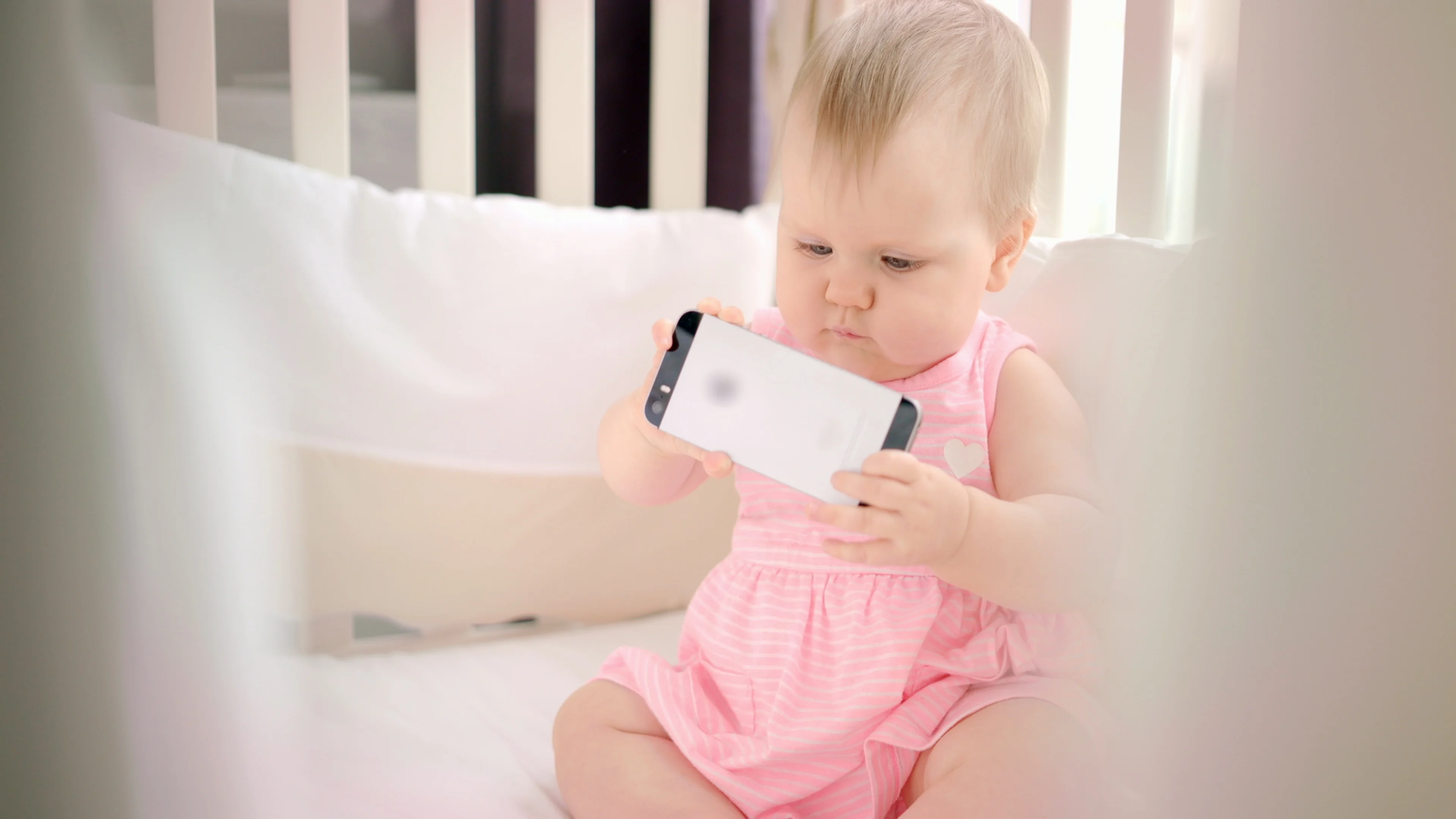 Little baby girl with phone in crib. Infant watching cartoon on mobile phone