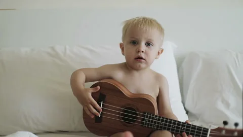 A little blond boy looks at the camera and plays ukulele. Stock Footage 94581278