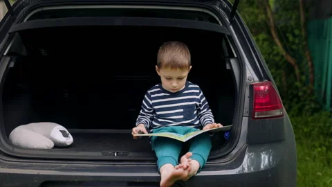 A little blond boy is sitting in the trunk of a car and looking at a book Video stock 196806409