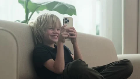 Little blonde boy using looking playing phone and sitting home on couch, holding Видео 231315788