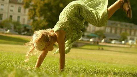 Little blonde girl performs handspring on a green park lawn on a sunny summer Stock Photos