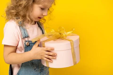 Little blonde girl on a yellow background holds a gift in a pink round box with Stock Photos