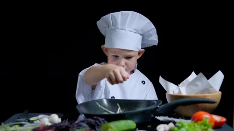 Little boy is adding a salt to the dish, wearing chefs suit and cap. Black Stock Footage 92555623