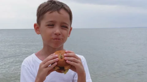 A little boy bites a hamburger. A happy child eats in nature, enjoying a Stock Footage 165652555