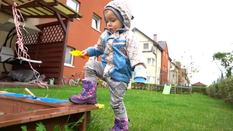 A little boy in boots and a jacket plays with a sandbox near the house. Children Stock Footage 146540205