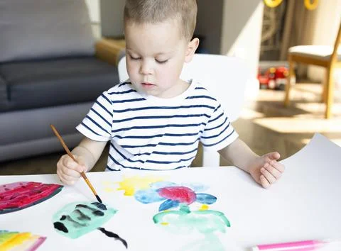 A little boy draws patterns on a white sheet with watercolor paints. Stock Photos