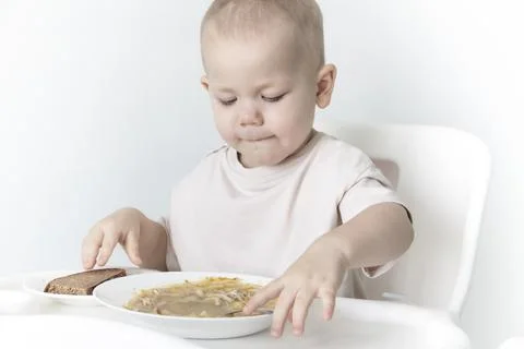 A little boy eats soup with bread on his own in a highchair against the backg Stock Photos