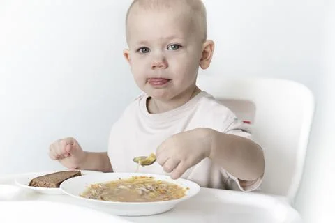 A little boy eats soup with bread on his own in a highchair against the backg Stock Photos