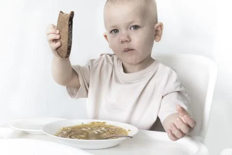 A little boy eats soup with bread on his own in a highchair against the backg Stock Photos