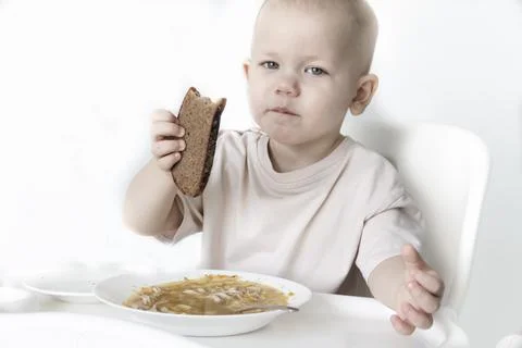 A little boy eats soup with bread on his own in a highchair against the backg Stock Photos