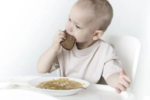 A little boy eats soup with bread on his own in a highchair against the backg Stock Photos