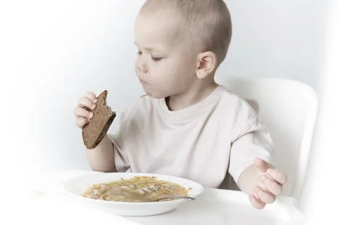A little boy eats soup with bread on his own in a highchair against the backg Stock Photos