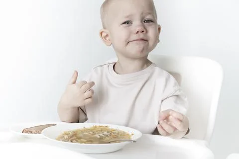 A little boy eats soup with bread on his own in a highchair against the backg Stock Photos