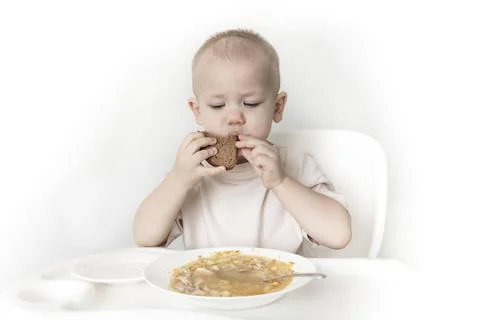 A little boy eats soup with bread on his own in a highchair against the backg Stock Photos