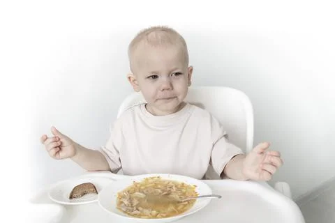 A little boy eats soup with bread on his own in a highchair against the backg Stock Photos