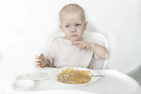 A little boy eats soup with bread on his own in a highchair against the backg Stock Photos