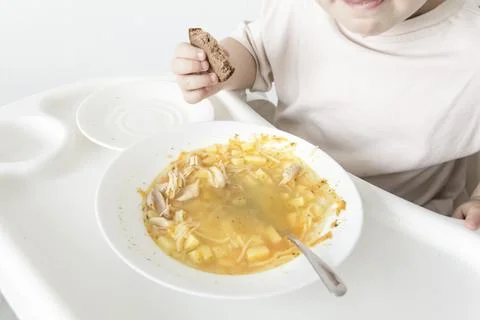 A little boy eats soup with bread on his own in a highchair against the backg Stock Photos