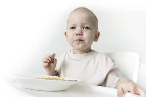 A little boy eats soup with bread on his own in a highchair against the backg Stock Photos