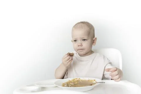 A little boy eats soup with bread on his own in a highchair against the backg Stock Photos