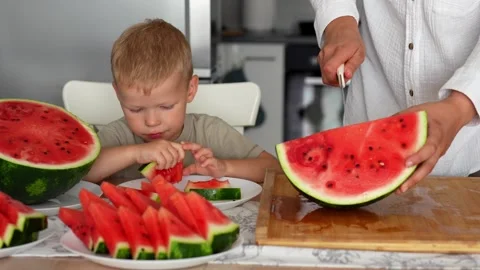 Little boy eats watermelon at the kitchen table at home while his mom Stock Footage 290982068