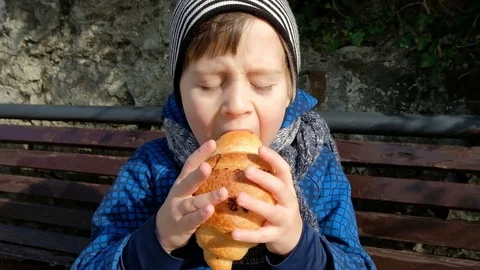 Little boy enjoy eating large croissant sitting on bench outdoor Stock Footage 74256953