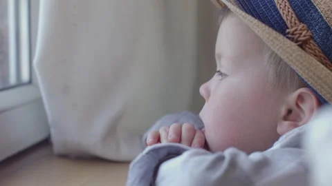 Little boy in hat sits at window, looks sadly out window, watches life outside Video stock 242604140